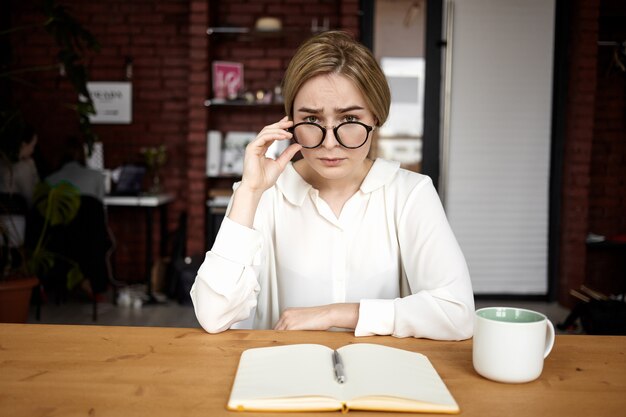 Ernstige mooie jonge vrouwelijke werkgever dragen wit overhemd en bril uitvoeren van sollicitatiegesprek zittend op kantoor met open dagboek en kopje koffie op houten bureau. Bedrijf en carrière