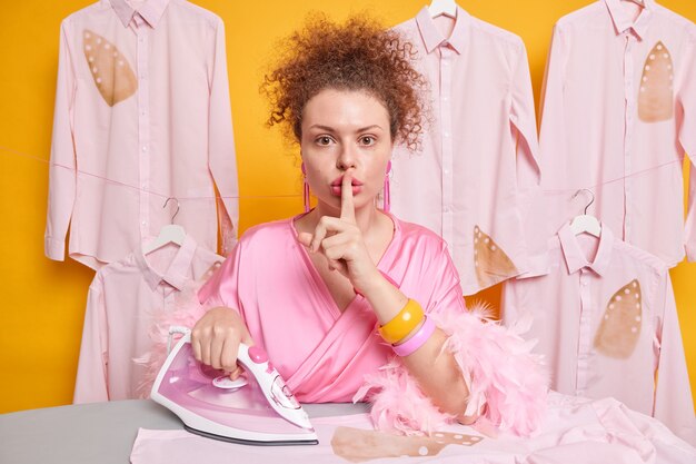 Ernstige mysterieuze huisvrouw met krullend haar maakt stiltegebaar en vraagt niemand te vertellen dat ze haar shirt heeft verbrand tijdens het strijken, omdat gebrek aan ervaring een kamerjas draagt die over een gele muur is geïsoleerd.