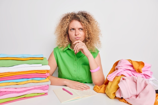 Ernstige nadenkende gekrulde vrouw verzamelt kleding voor donatie en maakt aantekeningen in dagboek omringd door opgevouwen en uitgevouwen wasgoed