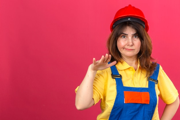 Gratis foto ernstige ogende bouwer vrouw in bouw uniform en veiligheidshelm probeert kalmeren vriend die ongeveer fout maken staande met opgeheven palm vragen om te stoppen over geïsoleerde roze muur met agent