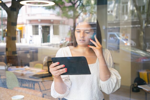 Ernstige onderneemster die op cel spreekt, tablet gebruikt en scherm bekijkt