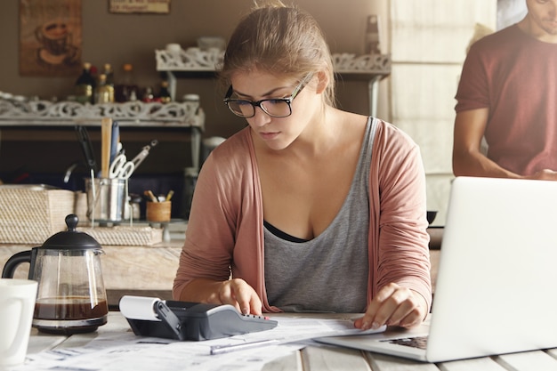 Ernstige ongelukkig jonge vrouw in glazen zitten aan de keukentafel met opengeklapte laptop pc en rekenmachine op het tijdens het berekenen van de financiën. Huisvrouw die elektronische apparaten gebruikt voor het online betalen van energierekeningen