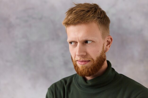 Ernstige ongeschoren jongeman in stijlvolle donkergroene coltrui die argwaan uitdrukt, wegkijkend van onder zijn wenkbrauwen. Knappe man met baard poseren, met een intense blik