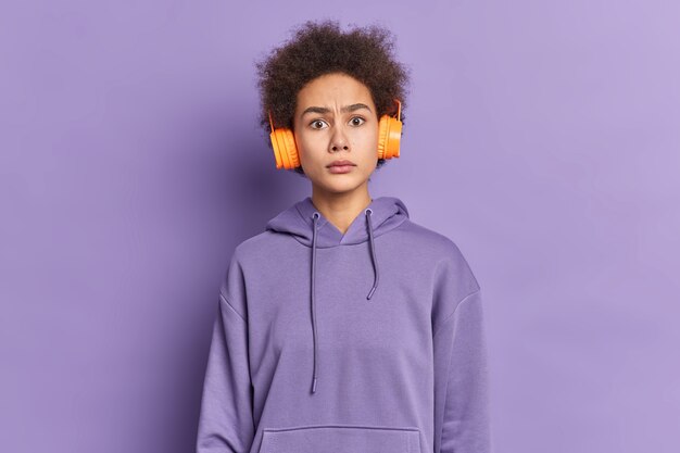 Ernstige ontevreden Afro-Amerikaanse vrouw met krullend haar ziet er verbaasd uit, draagt een stereohoofdtelefoon, luistert naar muziek terwijl ze op straat loopt, gekleed in een hoodie.