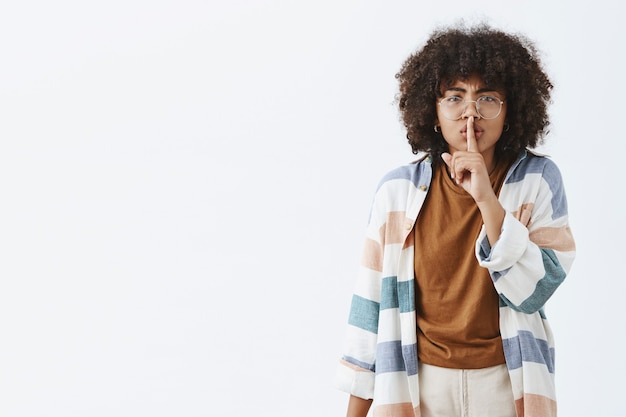 Ernstige ontevreden Afro-Amerikaanse vrouwelijke docent met afro kapsel in trendy kleding zegt shh tonen stil gebaar met wijsvinger over mond veeleisende stilte tijdens les