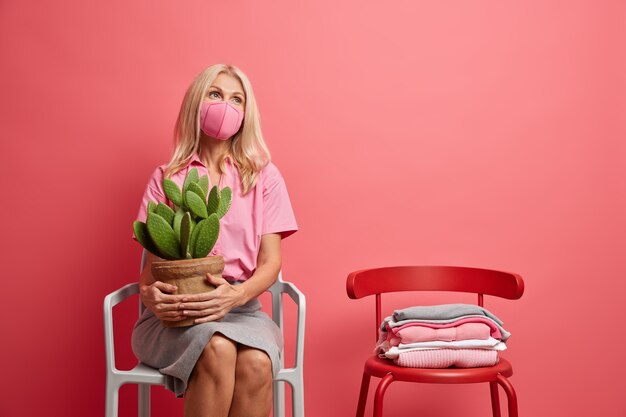 Ernstige peinzende vrouw van middelbare leeftijd draagt beschermend gezichtsmasker denkt aan veiligheid tijdens pandemie houdt ingemaakte cactus zit alleen op stoel blijft thuis tijdens quarantaineperiode. Covid 19 sociale afstand nemen