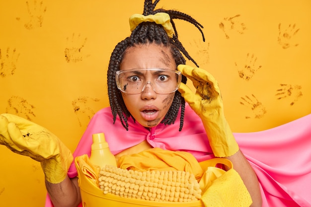 Ernstige slordige vrouw met gekamde dreadlocks die vies is na het opruimen van de kamer kijkt aandachtig door een transparante bril draagt een superheldenkostuum geïsoleerd over gele muur