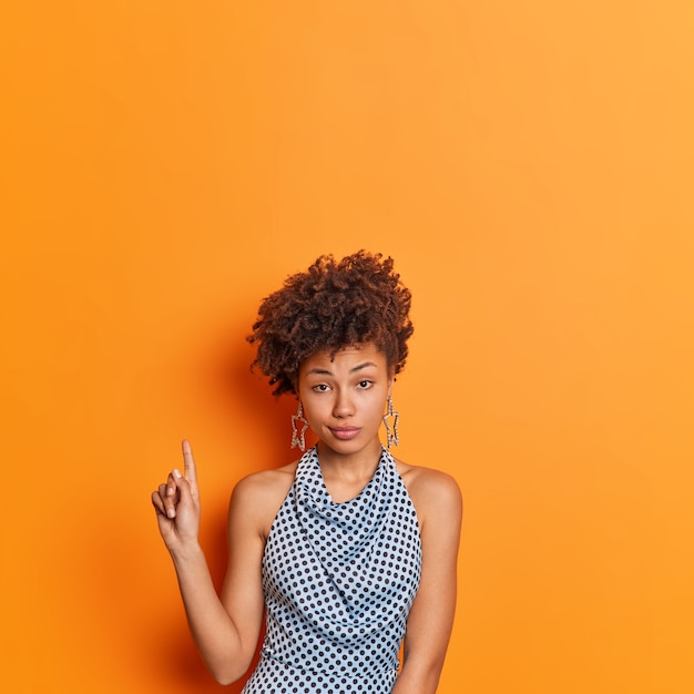 Ernstige stijlvolle jonge Afro-Amerikaanse vrouw in modieuze polka dot kleding geeft boven op kopie ruimte aanbeveling poses geeft tegen levendige oranje achtergrond. Kijk maar naar deze aanbieding