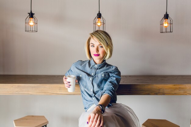 Ernstige stijlvolle meisje met blond haar en roze lippen, zittend in een coffeeshop met houten stoelen en tafel. Ze houdt een kopje koffie vast