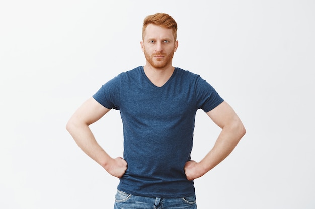 Ernstige strenge en knappe roodharige man in blauw t-shirt, handen vasthouden aan de taille en staren, iemand schelden of aanwijzingen geven, bazig zijn