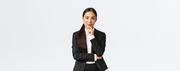 Ernstige tevreden Aziatische zakenvrouw heeft een interessant idee om de kin aan te raken en sluw naar de camera te kijken die doordacht denkt terwijl ze in pak staat op witte achtergrond