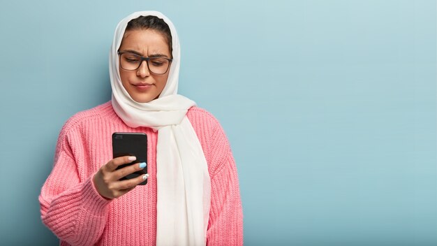 Ernstige verbaasde vrouw met een specifiek uiterlijk, die erg religieus is, draagt een witte sjaal, kijkt met een verbijsterde uitdrukking naar slimme telefoon, leest een vreemd bericht, geïsoleerd op blauwe muur, lege ruimte