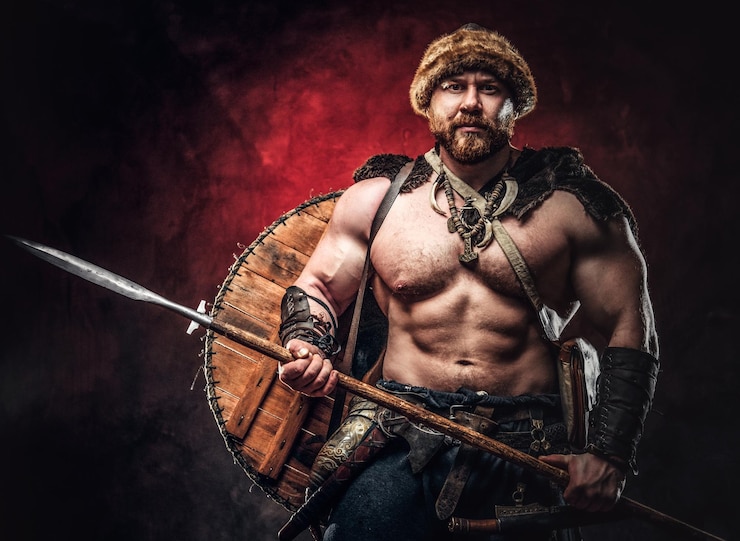 Gratis Foto ernstige viking gekleed in een licht harnas met een schild achter zijn rug houdt een speer vast. poseren op een donkere achtergrond met rood licht