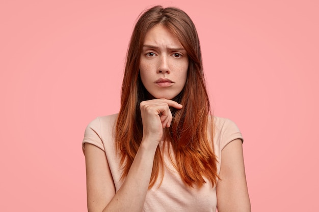 Ernstige vrouw met sproeten huid, hand onder de kin, fronsend gezicht, nonchalant gekleed, geïsoleerd op roze achtergrond.