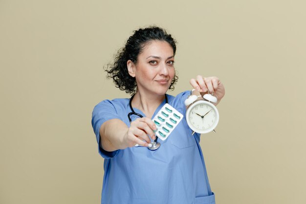 Ernstige vrouwelijke arts van middelbare leeftijd, gekleed in uniform en stethoscoop om de nek, kijkend naar camera die wekker en pakje capsules uitrekt naar camera geïsoleerd op olijfachtergrond