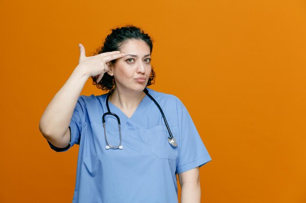 Ernstige vrouwelijke arts van middelbare leeftijd, gekleed in uniform en stethoscoop om haar nek, kijkend naar camera die zelfmoordgebaar maakt geïsoleerd op oranje achtergrond