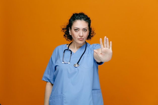 Ernstige vrouwelijke arts van middelbare leeftijd, gekleed in uniform en stethoscoop om haar nek, kijkend naar camera met stopgebaar geïsoleerd op oranje achtergrond