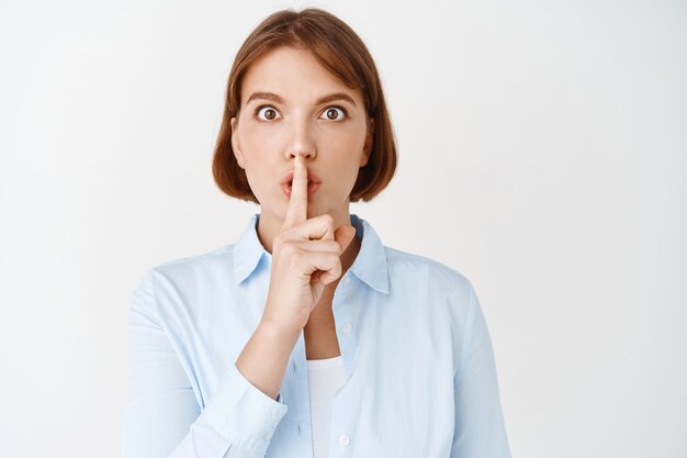 Ernstige vrouwelijke kantoordame zwijgt, drukt de vinger op de lippen en kijkt bezorgd, zeg dat je stil moet zijn, maak een gebaar, witte muur