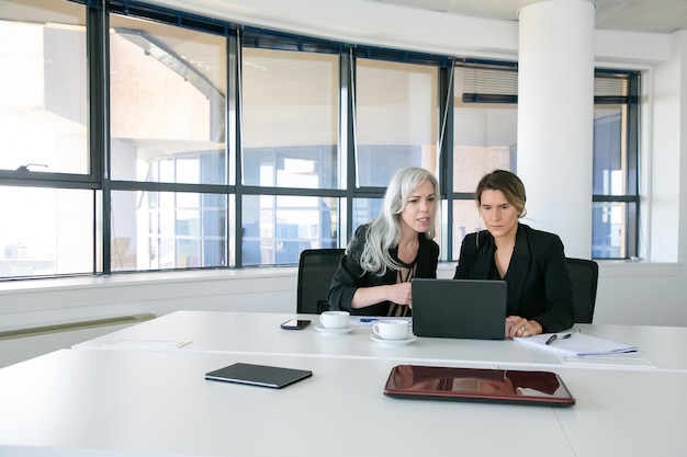 Ernstige zakelijke dames kijken en bespreken van inhoud op laptop zittend aan tafel met kopjes koffie in de vergaderruimte. Breed schot. Teamwork en communicatieconcept