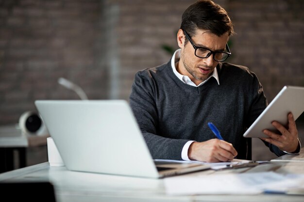 Ernstige zakenman die aan touchpad werkt en rapporten op het papier op kantoor schrijft