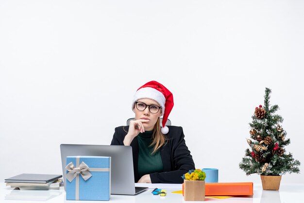 Ernstige zakenvrouw met haar kerstman hoed zittend aan een tafel met een kerstboom en een cadeau erop en gericht op iets zorgvuldig op witte achtergrond