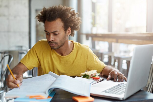 Ernstige zelfverzekerde jonge donkere leraar in vrijetijdskleding bezig met onderwijsplan, notities maken met pen, zittend aan kantine met schoolboeken, schrijfboeken, notebook pc en sandwich op tafel