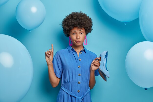 Ernstige zelfverzekerde vrouw punten hierboven nodigt je uit om naar boven te gaan, houdt nieuwe schoenen op hoge hakken, jurken in modieuze outfit, probeert kleren om uit te gaan, poseert tegen blauwe muur met ballonnen