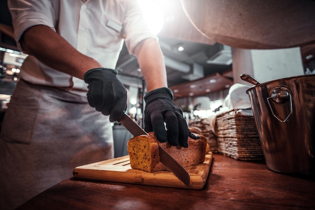 Ervaren bakker in beschermende handschoenen snijdt brood voor dagelijks ontbijt in restaurant.