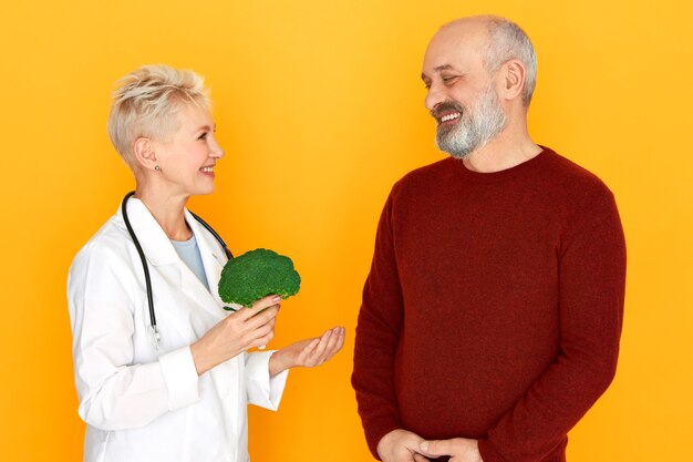 Ervaren vrouwelijke arts van middelbare leeftijd die broccoli in haar handen houdt, praat over de voordelen van gezonde biologische voeding aan bebaarde oudere mannelijke patiënt