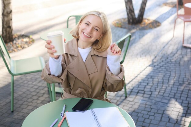 Gratis foto eten en drinken concept jonge gelukkige vrouw zitten in een café genieten van een heldere dag buiten verhogen kopje koffie