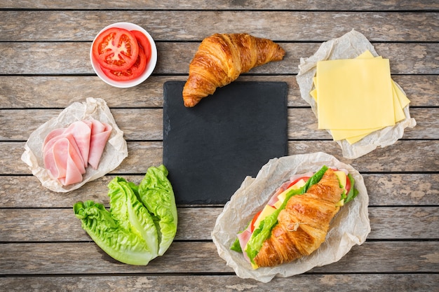 Eten, ontbijt, ochtend en lunch, diy, doe het zelf concept. verse croissantsandwich met ingrediënten, ham, kaas, sla en tomaat op een houten tafel. platliggende achtergrond