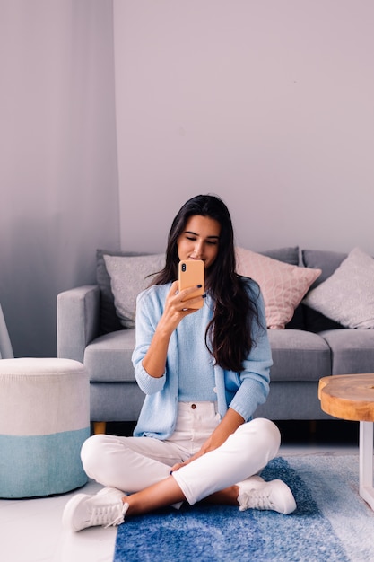 Europese fit brunette fashion blogger vrouw zit op de vloer in de woonkamer in de buurt van bank met telefoon