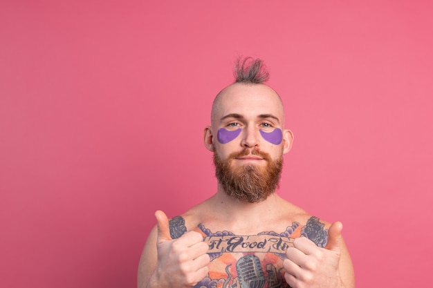 Europese knappe bebaarde getatoeëerde topless man met paarse eye patches masker poseren voor camera op roze