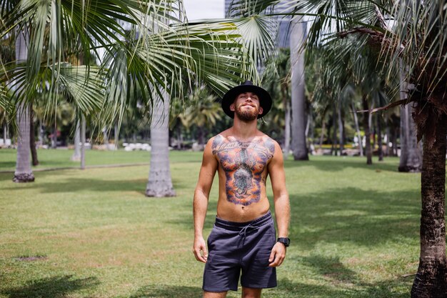 Europese knappe getatoeëerde sterke bebaarde man in klassieke hoed topless op veld in tropisch park