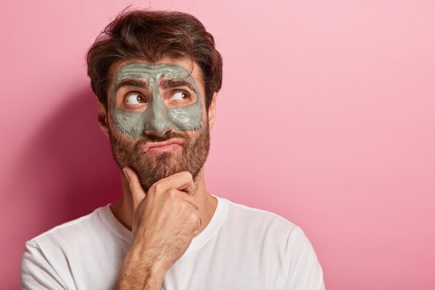 Europese man houdt kin vast, tuit lippen, heeft een bedachtzame uitdrukking, dikke haren, kijkt opzij, past kleimasker op gezicht, draagt een wit t-shirt verfrist de huid, modellen over roze muur. Mannelijke cosmetologie