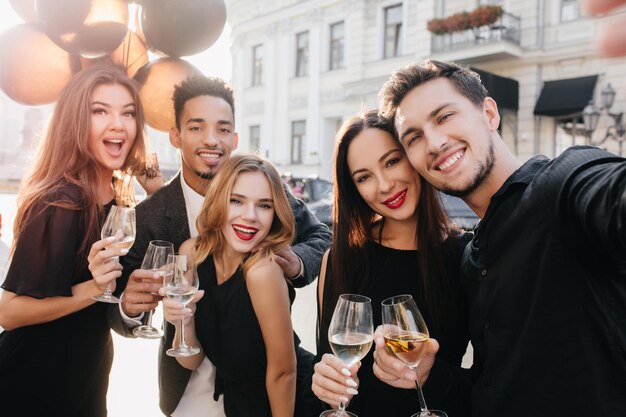 Europese man met schattige glimlach die selfie maakt tijdens straatfeest op zonnige dag. Aanbiddelijke blanke vrouw die met feestballonnen zwaait terwijl ze wijnglas vasthoudt op de achtergrond van de wazige stad.