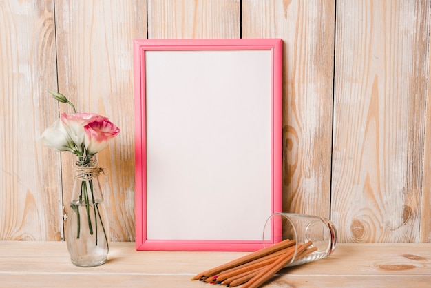 Eustoma in glazen vaas; kleurpotloden en witte fotolijst met roze rand op houten tafel