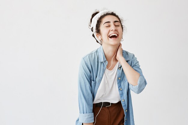 Expressief gelukkig emotioneel meisje met donker en krullend haar in knot, luistert naar muziek in oortelefoons, geniet van aangename melodieën, heeft een goed humeur, ontspant binnenshuis. Mensen, vrije tijd, levensstijl, schoonheid.