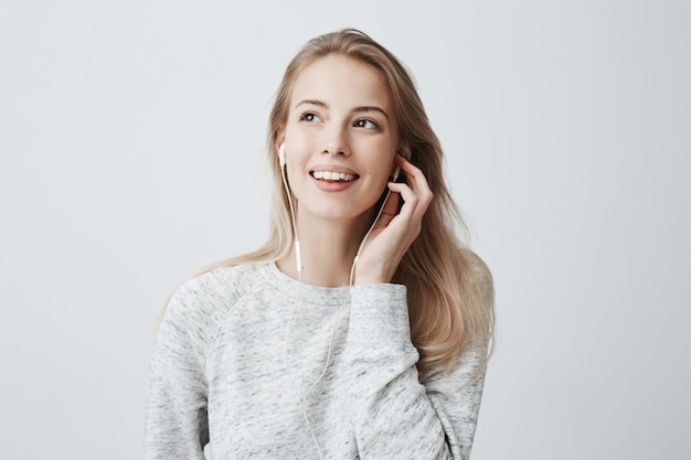 Expressieve gelukkige jonge blanke vrouw draagt los geverfd blond haar, luistert naar muziek in oortelefoons, geniet van aangename melodieën, heeft een goed humeur.