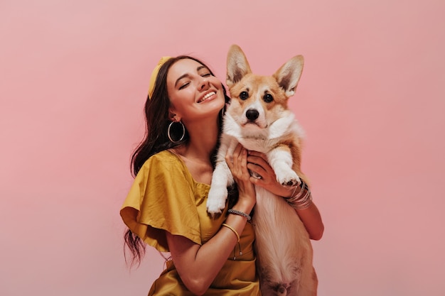 Exquise meisje met lang golvend haar in ronde oorbellen, trendy bandana en gele stijlvolle jurk poseren met gesloten ogen en glimlachen