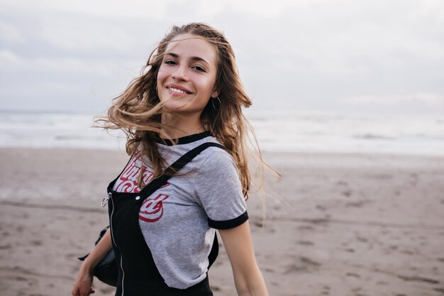 Extatisch bruinharige meisje springen op het strand in bewolkte zomerdag. Foto van aantrekkelijk wit vrouwelijk model poseren op aard.