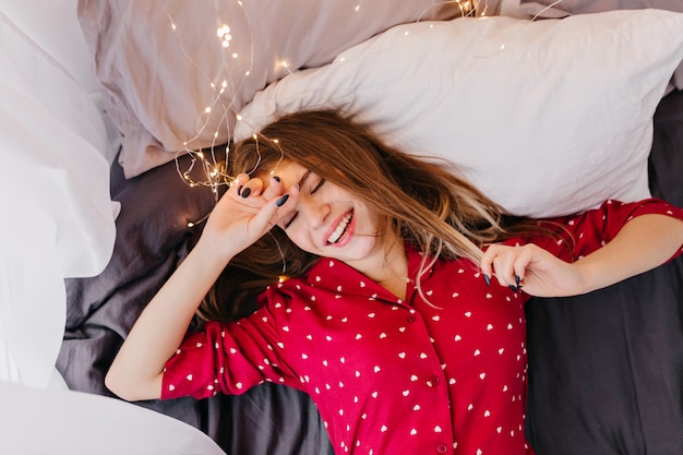 Extatisch kaukasisch meisje met bruin haar dat in bed ligt en glimlacht. lachend vrouwelijk model in rode nachtkleding genieten van goedemorgen.