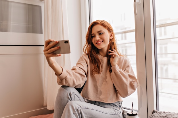 Extatisch meisje met donker golvend haar selfie maken. Mooie gember vrouw zitten in haar kamer met smartphone.