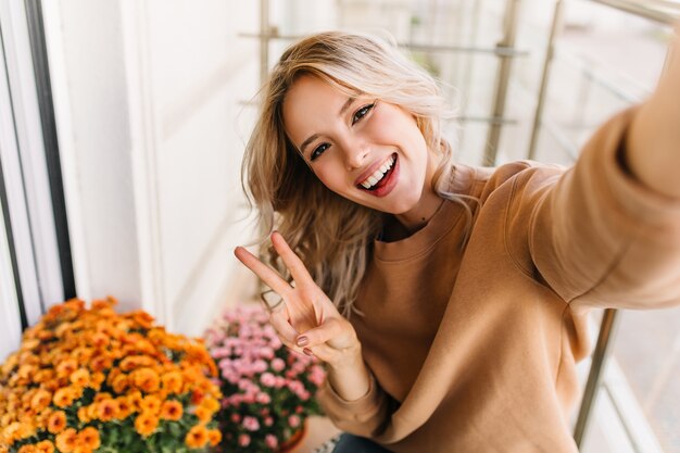 Extatische caucaisan vrouw selfie met vredesteken maken. Elegant krullend meisje poseren met oranje bloemen.