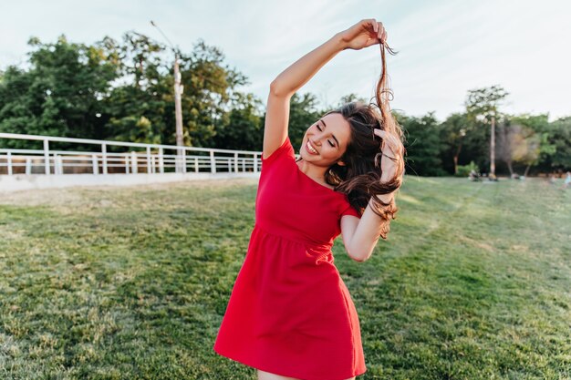 Extatische jonge dame in zomerkleding spelen met haar haren tijdens fotoshoot in park. Buiten schot van schattig meisje in een rode jurk met plezier in weekend.