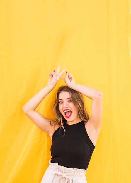 Gratis foto extatische jonge vrouw die vredesteken tonen die zich tegen gele achtergrond bevinden