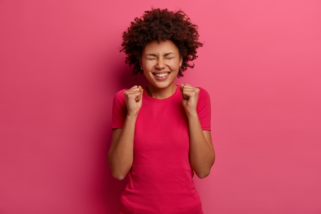 Extatische vrolijke vrouw maakt vuist hobbel, voelt zich erg gelukkig, verheugt zich op succes en triomf, viert veel geld in loterijen, sluit ogen, gekleed in roze kleren, poseert binnen. Overwinning behaald