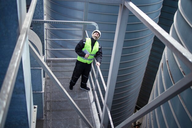 Fabrieksarbeider staande op metalen platform tussen industriële opslagtanks en op zoek naar visuele inspectie van silo's voedselproductie