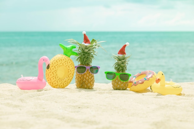 Familie van grappige aantrekkelijke ananassen in stijlvolle zonnebril op het zand tegen turquoise zee. kerstmutsen dragen. kerstmis en nieuwjaar vakantie concept op tropisch strand. vakantie met het gezin. helder