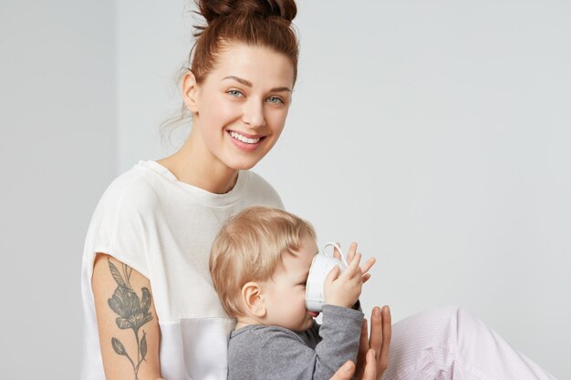 Familieportret van glimlachende moderne moeder en haar baby op witte muur. Gelukkig moeder in witte pyjama zittend met haar zoon. Leuke babyjongen in grijs shirt consumptiemelk uit stijlvolle witte beker.